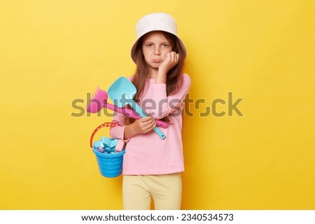 Bored sad little girl wearing casual clothing and panama holding sandbox toys and bucket isolated over yellow background feeling disinterest at playground posing with rake and scoop.