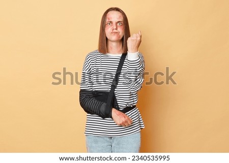 Angry sick woman with scratches and bruises on her face standing isolated over beige background showing fist after being beating revenge. Royalty-Free Stock Photo #2340535995