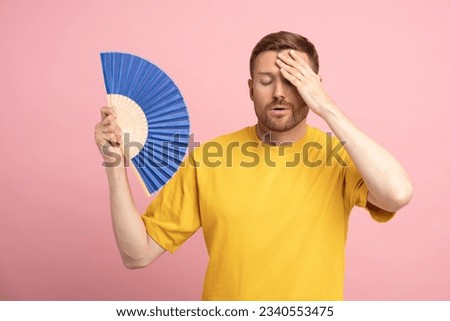 Too hot. Sweaty tired middle aged man touching forehead using paper fan suffer from heat, feels sluggish. Displeased guy cooling in hot summer weather, isolated on studio pink background. Overheating Royalty-Free Stock Photo #2340553475
