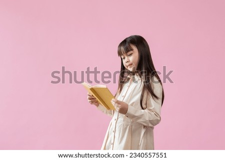 A cute young Asian elementary school girl student is focused on reading a book while standing against an isolated pink studio background. education, children, kids, leisure and hobby