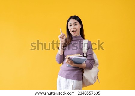 A beautiful and cheerful young Asian female college student with a backpack is carrying her books and pointing her finger up while standing against an isolated yellow background.