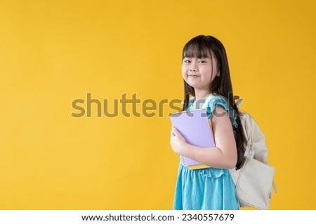 An adorable young Asian girl with books in her hand and a backpack is standing against an isolated yellow studio background with copy space. kindergarten, kids, children, elementary school student