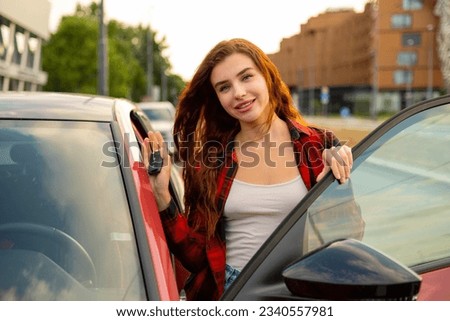 Red-haired girl sporting a trendy plaid shirt stands beside a flashy red car, her hands clutching the keys Royalty-Free Stock Photo #2340557981