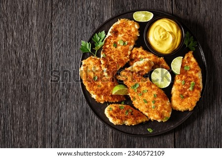 fried chicken cutlets under breadcrumbs and parmesan cheese crust on black plate with mustard and lime on dark wooden table, horizontal view from above, flat lay, copy space Royalty-Free Stock Photo #2340572109