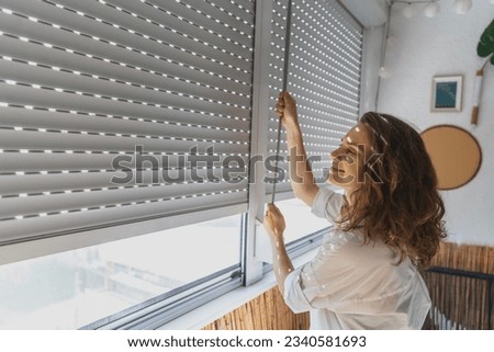 Young woman closing roller shutters on the balcony on a summer day Royalty-Free Stock Photo #2340581693