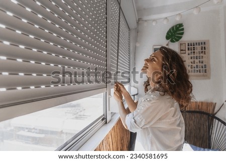 Young woman closing roller shutters on the balcony on a summer day Royalty-Free Stock Photo #2340581695