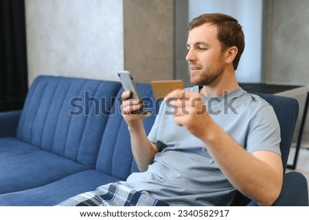 A happy smiling caucasian guy, in casual stylish clothes, sits on the couch at home, holds a phone and a banking card in his hands, makes online payment for purchases, enters a card number.