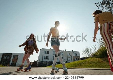 Young fit woman in roller skates listening to music in headphones and skating in front of camera among two friends against sunshine