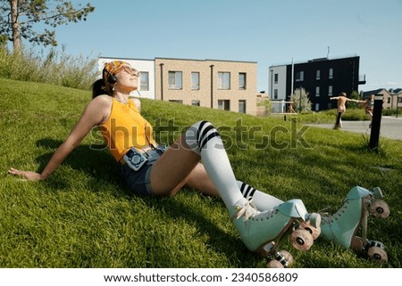 Young relaxed woman with walkman and headphones listening to music while sitting on green lawn in urban environment and enjoying break Royalty-Free Stock Photo #2340586809