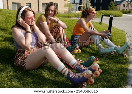 Young woman with headphones listening to music while sitting on green lawn and having rest with her friends after roller skating in the city Royalty-Free Stock Photo #2340586815