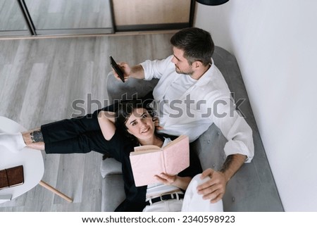 From above smiling young woman with book looking at camera while lying on knees of boyfriend browsing smartphone on sofa Royalty-Free Stock Photo #2340598923