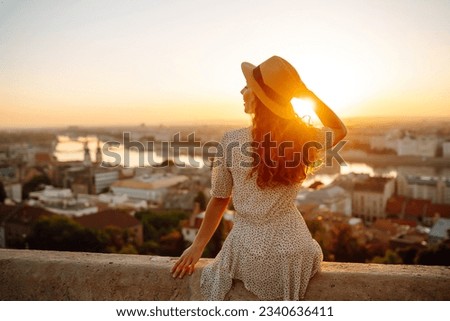 Happy traveler in a stylish dress and hat enjoys the sunrise or sunset with stunning views of the city. Back view. Lifestyle, travel, tourism, nature, active lifestyle. Royalty-Free Stock Photo #2340636411