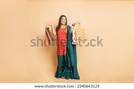 Portrait of young beautiful indian woman wearing traditional outfit holding shopping bags standing isolated on beige studio background. Diwali celebration and festive sale concept.Full length. Royalty-Free Stock Photo #2340643129