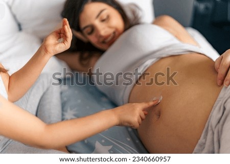A picture of a pregnant mom smiling while her daughter rubs lotion on her belly, spending quality time until the baby arrives