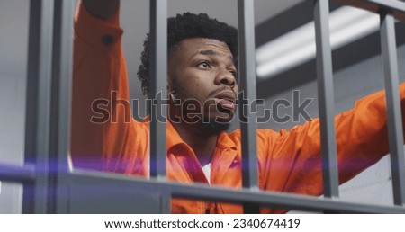 Sad African American man in orange uniform leans hands on prison cell bars and kneads neck. Depressed prisoner serves imprisonment term in jail. Murderer in correctional facility or detention center. Royalty-Free Stock Photo #2340674419