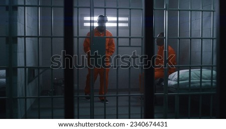 Two African American prisoners in orange uniforms talk. One man sits on bed, another walks in prison cell. Men serve imprisonment terms for crimes in jail, detention center or correctional facility. Royalty-Free Stock Photo #2340674431