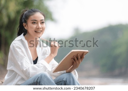 Photo of young attractive Asian woman writing on diary while sitting outdoors in the park, Writer sitting in garden, Female freelance are relaxing to writing story on jobs, beautiful Asian women