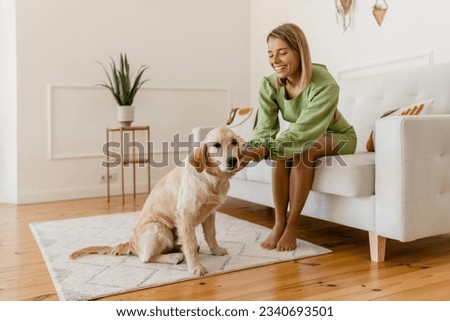 young stylish smiling woman in green summer dress in modern interior apartment home relaxing fashion style trend sitting on couch petting dog golden retriever breed Royalty-Free Stock Photo #2340693501