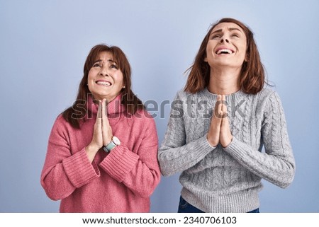 Mother and daughter standing over blue background begging and praying with hands together with hope expression on face very emotional and worried. begging. 