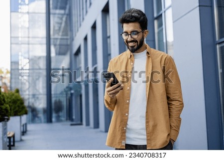 Young successful businessman walking in the city outside office building, hispanic smiling happy with achievement results, holding phone, using online application outdoors. Royalty-Free Stock Photo #2340708317