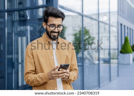 Young successful businessman walking in the city outside office building, hispanic smiling happy with achievement results, holding phone, using online application outdoors. Royalty-Free Stock Photo #2340708319