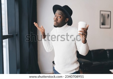 Confused African American male in hat standing with cup of hot drink and looking at camera while shrugging shoulders in misunderstand in living room Royalty-Free Stock Photo #2340708951