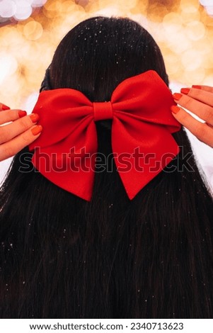 Female hands in a knitted winter sweater holding a red bow in black hair in a festive New Year atmosphere with magic bokeh lights