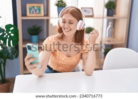 Young redhead woman using smartphone doing video call sitting on the table screaming proud, celebrating victory and success very excited with raised arm 