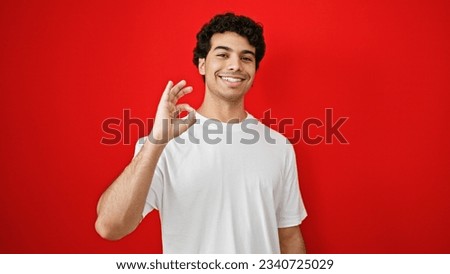 Young latin man doing ok gesture smiling over isolated red background