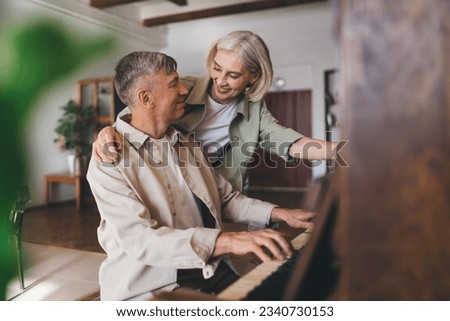 Happy aged female in casual clothes hugging man playing melody on piano during weekend together at home looking at each other and smiling Royalty-Free Stock Photo #2340730153