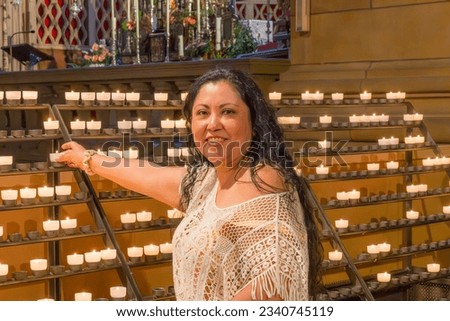 Smiling Mexican woman lighting a candle in a church, looking at camera, chandelier with tea light candles in background, long wavy black hair, see-through knitted blouse Royalty-Free Stock Photo #2340745119