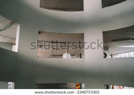 Unemotional young man in glasses and white t shirt standing in spacious round shaped concrete parking building with geometric windows located near urban resident house