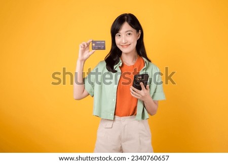 Excited young Asian woman 30s, dressed in orange shirt and green jumper, uses smartphone to show credit card on yellow studio background. Online shopping from mobile concept.