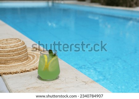 Close up shot of broad brim straw hat and a glass of ice cold lemonade with mint on the edge of the pool deck. Copy space, background.