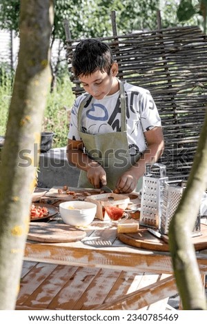 A boy in an olive-colored apron makes pizza enthusiastically. Cuts sausages into small pieces. The boy on the background of trees in the garden.
