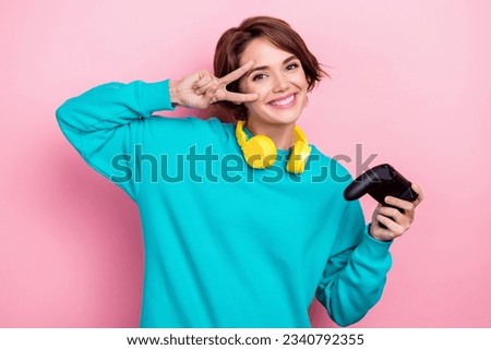 Photo portrait of pretty young girl hold joystick show v-sign headphones dressed stylish blue outfit isolated on pink color background