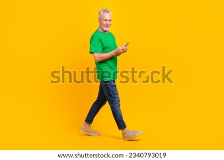 Full size profile photo of cheerful aged man walking hold smart phone typing blog post isolated on yellow color background