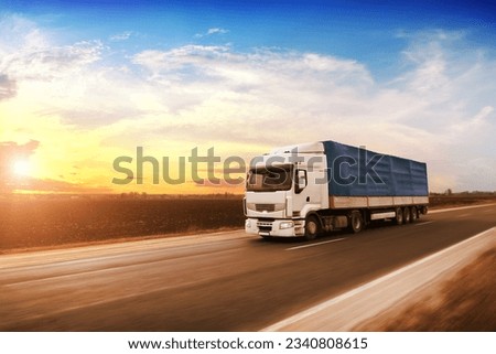 Big white truck with a blue trailer with blank space for text on a countryside road in motion against an evening  sky with a sunset Royalty-Free Stock Photo #2340808615