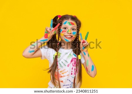 A happy little girl painted with colorful paints. A child with a face smeared with paints, is fond of drawing and shows bunnies from his fingers. Yellow isolated background. Royalty-Free Stock Photo #2340811251
