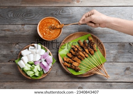 Overhead view people holding chicken satay dipping peanut sauce on wooden dining table, one of famous local dishes.