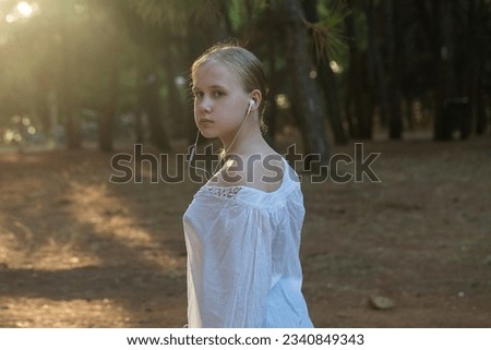 Nice teenage girl in white shirt listen to music in headphones on the background of forest and sunlight