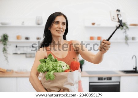 Close up of adult in terracotta tank top taking self-picture via smartphone boom arm while carrying groceries in bag. Beautiful lady encouraging to healthy well-being via eating habits and training.