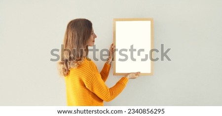 Woman decorating interior, hanging white blank photo frame, wooden picture, artwork mockup on wall in new house