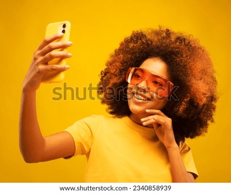 Young dark skinned  woman with Afro hairstyle, holds mobile phone, makes video call, dressed in casual wear and orange sunglasses, isolated over yellow studio wall.
