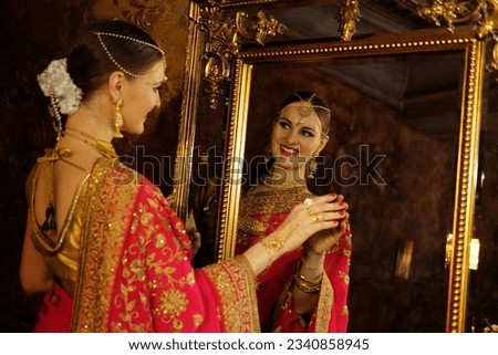 Female model Hindu Bride in saree, wearing gold and jasmine flower garlands in the hair looks in the mirror and touches his reflection, back view. Royalty-Free Stock Photo #2340858945