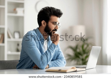 Pensive Young Indian Male Freelancer In Eyeglasses Working With Laptop At Home Office, Handsome Eastern Man Looking At Computer Screen And Touching Chin, Thinking About Business Project, Free Space