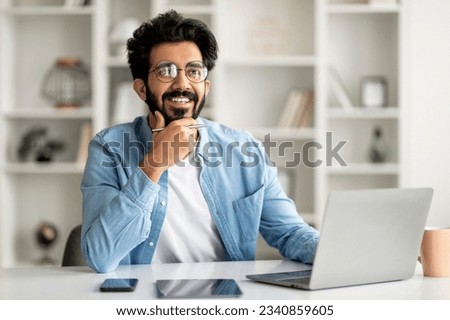 Dreamy Young Indian Man Sitting At Desk With Laptop In Home Office And Looking Away, Smiling Eastern Male Freelancer Thinking About Business Plans Or Having Inspiration Moments, Touching Chin Royalty-Free Stock Photo #2340859605