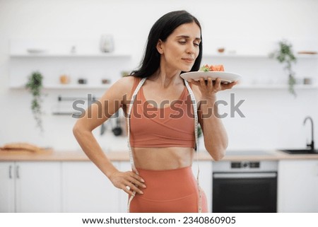 Pretty brunette woman enjoying smell of healthy organic salad while standing in kitchen after regular workout. Athletic female with prefect body shape consuming low calorie food to avoid weight gain. Royalty-Free Stock Photo #2340860905