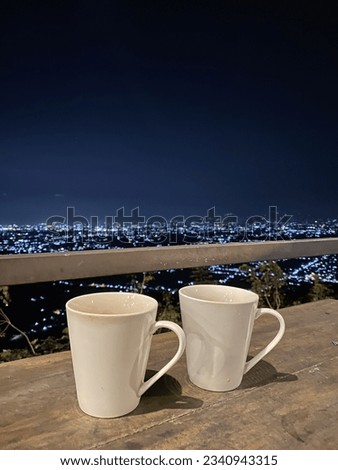 In a high mountain range, two coffee mugs rest gracefully, bathed in the warm glow of the rising sun. Majestic mountains frame city lights twinkling in the distance, harmoniously blending nature.
