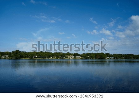 This is a photo of a lake and houses in the distance, with blue sky, white clouds, and calm water.                               
